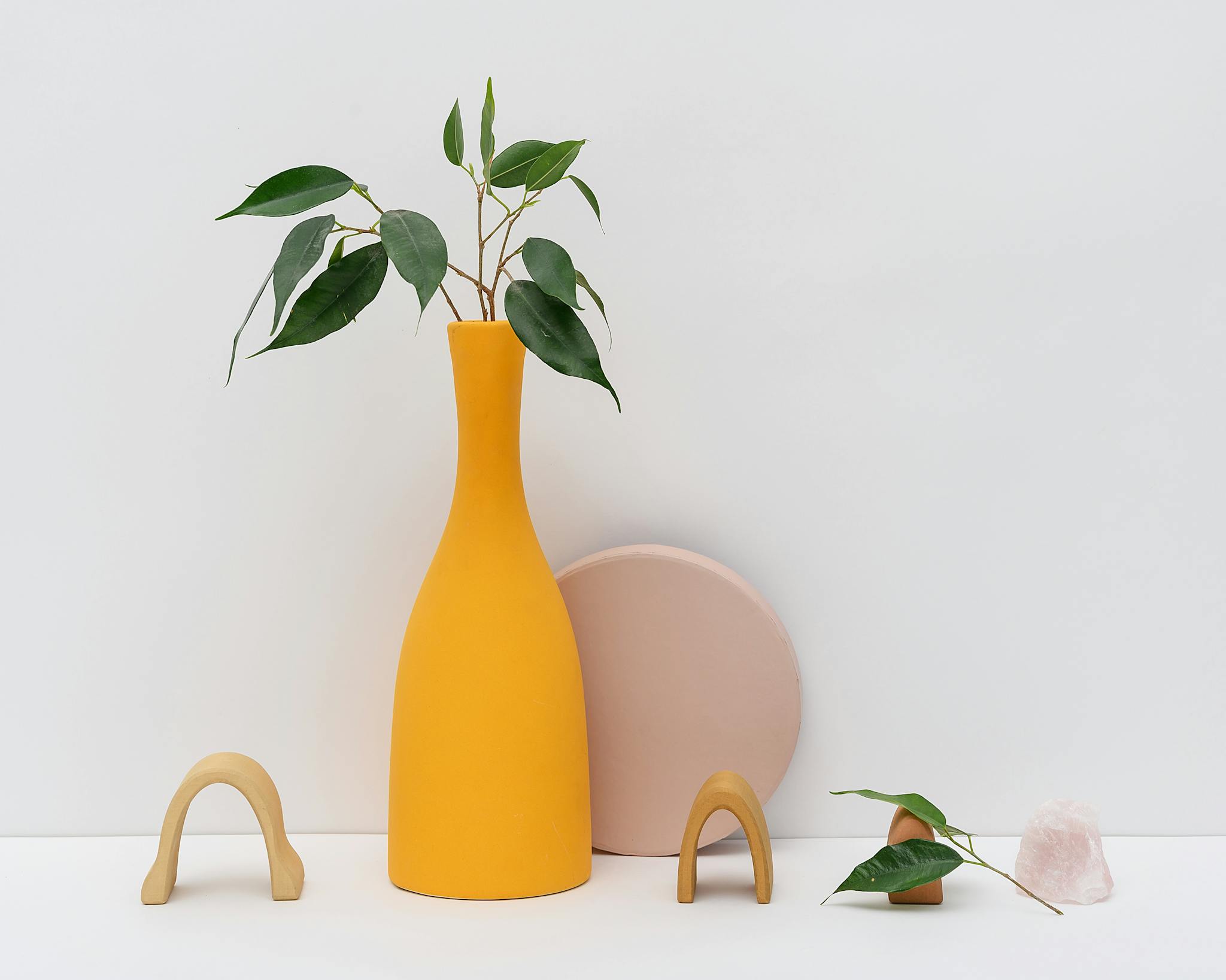 Modern still life featuring a yellow vase and green leaves in a minimal setting.